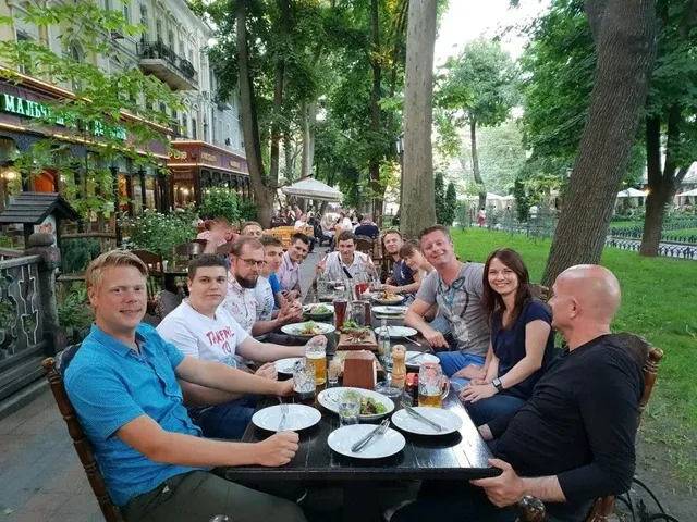 

Friends sharing food & smiles at an outdoor table covered w/ plants &